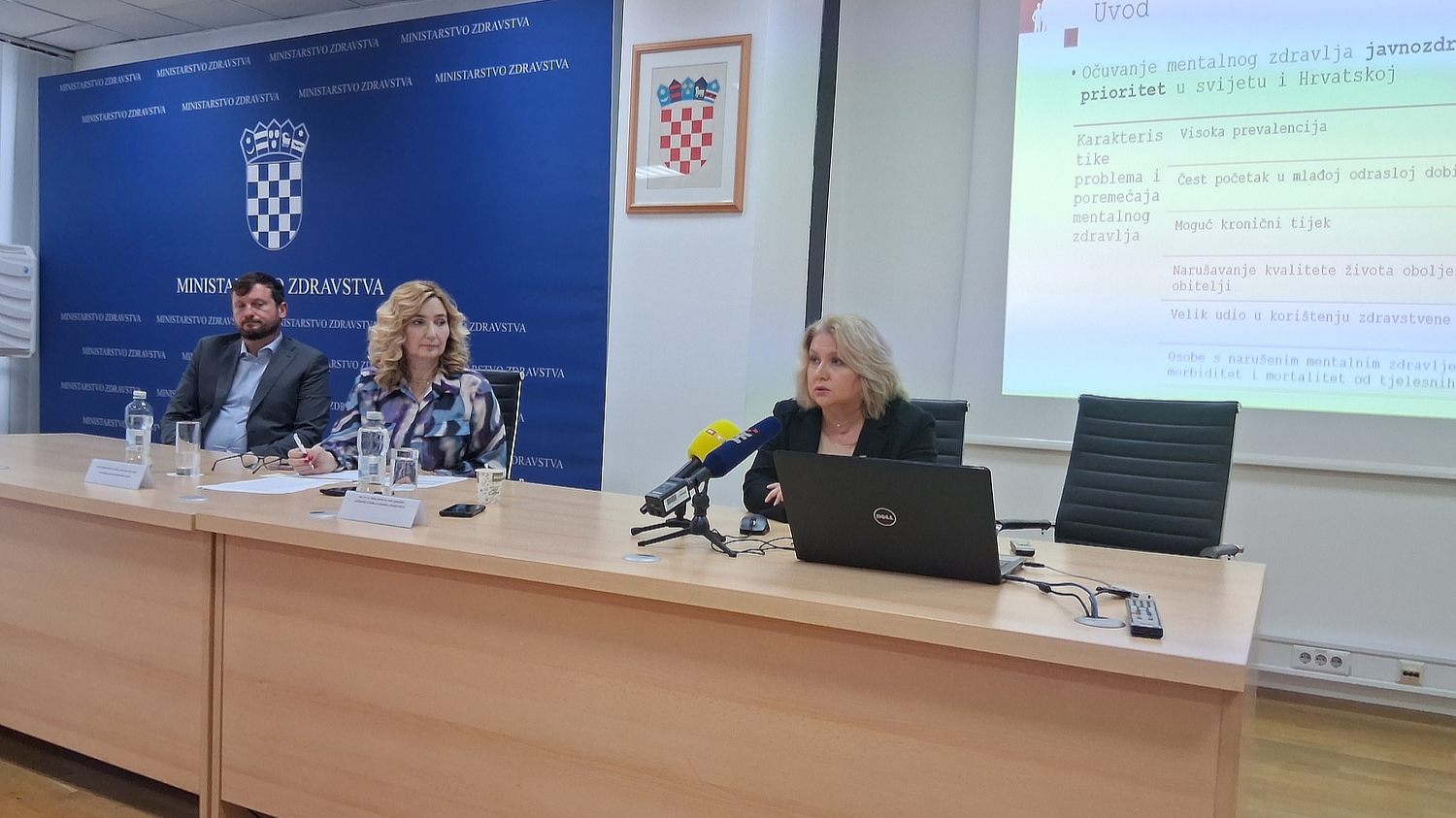 Tijekom konferencije u Ministarstvu zdravstva tri osobe sjede za stolom ispred plavog zida s državnim grbom, dok govornica iznosi podatke o očuvanju mentalnog zdravlja uz prezentaciju projiciranu na zidu.