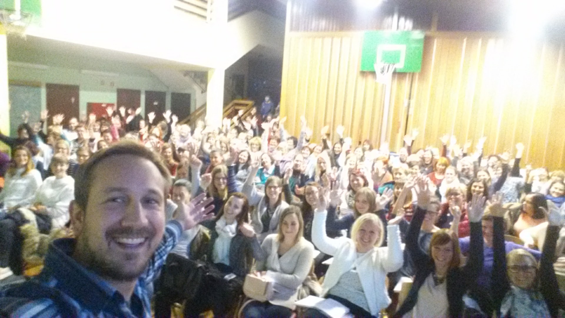 Na slici je prikazana velika skupina ljudi okupljena u prostoru koji izgleda kao višenamjenska dvorana – moguće školska sportska dvorana, s obzirom na drvene obloge na zidovima i košarkaški obruč u pozadini. Svi sjede i djeluju vedro, mnogi podižu ruke u znak uzbuđenja ili sudjelovanja. Atmosfera je topla i energična, puna zajedništva i pozitivne interakcije. U prvom planu, osoba snima selfie s grupom iza sebe – široko se smiješi, a njezin izraz lica odražava radost i ponos. Cijela scena odiše osjećajem zajedništva, angažiranosti i slavlja, kao da je riječ o radionici, predavanju ili posebnom događaju koji okuplja ljude s istim ciljem.