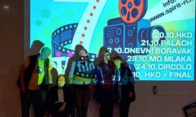 Fotografija prikazuje grupu od šest osoba koje stoje ispred svijetlog projekcijskog platna na kojem se prikazuje promotivni vizual filmskog događanja pod nazivom "CIJELI TJEDAN FILM 1 PO 1". Platno je ispunjeno živopisnim grafičkim elementima poput filmske trake, koluta i velikog broja 7, što može upućivati na sedmo izdanje festivala ili simboličnu poveznicu s brojem sedam. U prvom planu, osobe poziraju za zajedničku fotografiju, a jedna osoba sjedi u invalidskim kolicima, što jasno pokazuje inkluzivni karakter događanja. Atmosfera je prijateljska i svečana, a raznolikost sudionika doprinosi dojmu zajedništva i otvorenosti.