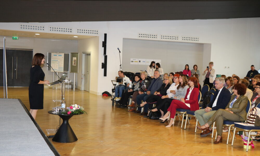 Na fotografiji je žena koja stoji za prozirnim govornim pultom i drži prezentaciju ili govor pred publikom u konferencijskoj dvorani. Publika, koju čine muškarci i žene, pažljivo sluša — neki bilježe, dok drugi koriste elektroničke uređaje. Prostor ima drveni pod, bijele zidove i moderan strop s ugrađenom rasvjetom, što stvara uredan i suvremen ambijent. Iza govornice se nalazi cvjetni aranžman na podu i stalak s mikrofonom, dok se na zidu u pozadini vide plakati ili informativni prikazi, što sugerira da je riječ o konferenciji, seminaru ili radionici. Cijela scena odiše profesionalnošću i ozbiljnošću, ali i toplinom — govornica djeluje samouvjereno i angažirano, a prisutnost cvijeća unosi dozu pažnje prema detaljima i estetskoj ugodi. Fotografija prenosi važnost dijeljenja znanja, javnog nastupa i međusobnog učenja u poslovnom, obrazovnom ili društvenom kontekstu.