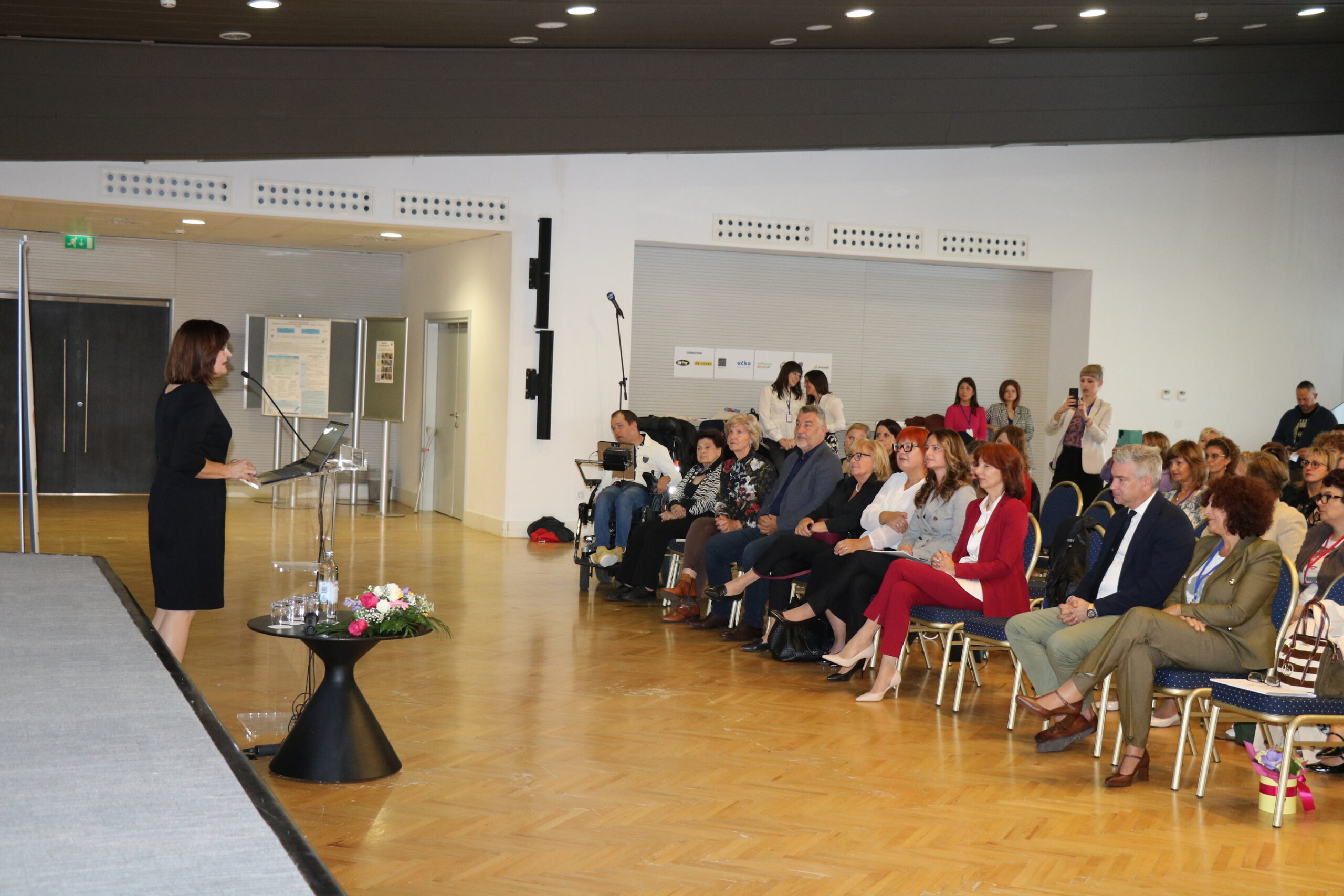 Na fotografiji je žena koja stoji za prozirnim govornim pultom i drži prezentaciju ili govor pred publikom u konferencijskoj dvorani. Publika, koju čine muškarci i žene, pažljivo sluša — neki bilježe, dok drugi koriste elektroničke uređaje. Prostor ima drveni pod, bijele zidove i moderan strop s ugrađenom rasvjetom, što stvara uredan i suvremen ambijent. Iza govornice se nalazi cvjetni aranžman na podu i stalak s mikrofonom, dok se na zidu u pozadini vide plakati ili informativni prikazi, što sugerira da je riječ o konferenciji, seminaru ili radionici. Cijela scena odiše profesionalnošću i ozbiljnošću, ali i toplinom — govornica djeluje samouvjereno i angažirano, a prisutnost cvijeća unosi dozu pažnje prema detaljima i estetskoj ugodi. Fotografija prenosi važnost dijeljenja znanja, javnog nastupa i međusobnog učenja u poslovnom, obrazovnom ili društvenom kontekstu.