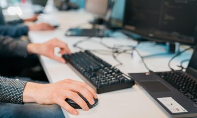Na fotografiji je prikazan krupni kadar radnog prostora osobe koja radi za računalom. Desna ruka koristi miš, dok lijeva tipka po tipkovnici, što sugerira aktivan rad u digitalnom okruženju. Na stolu se nalaze dva monitora s prikazom koda ili tehničkog teksta, što upućuje na programerski ili IT kontekst. Vidljive su i povezane žice te djelomično otvoreno prijenosno računalo s desne strane. Osoba nosi tamnu košulju s uzorkom dugih rukava. Cjelokupna scena odiše koncentracijom i tehničkom stručnošću, tipičnom za suvremeni radni prostor softverskog inženjera ili IT stručnjaka.