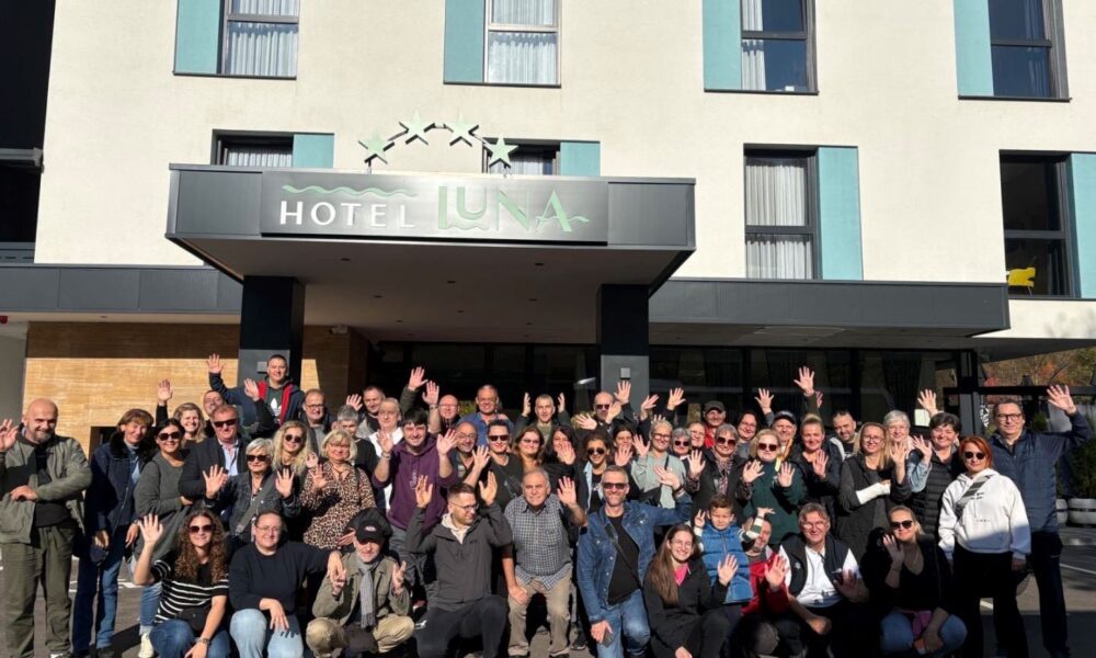 Ispred modernog hotela "LUNA", s bijelo-plavom fasadom i velikim prozorima, okupljena je velika skupina ljudi različitih dobi. Mnogi od njih nasmiješeno poziraju i mašu prema kameri, stvarajući dojam zajedništva i vedrine. Iznad ulaza jasno se ističe naziv hotela u stiliziranom fontu, ukrašen zvjezdicama koje dodaju notu elegancije. Atmosfera na fotografiji odiše prijateljstvom, kao da se radi o društvenom događaju, zajedničkom putovanju ili susretu punom pozitivne energije.