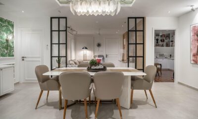 Fotografija prikazuje elegantno uređen, otvoreni prostor koji spaja blagovaonicu, dnevni boravak i radni kutak. U prvom planu nalazi se blagovaonski stol s mramornom površinom, okružen sa šest bež tapeciranih stolica s drvenim nogama. Na sredini stola smješten je dekorativni aranžman sa sukulentima, koji prostoru daje svježinu i prirodni ton. Iznad stola visi impresivan luster s višeslojnim staklenim ili kristalnim elementima, a zrcalni panel na stropu dodatno pojačava osjećaj dubine i sofisticiranosti. U pozadini se nalazi udoban dnevni boravak s bijelim kaučem, podnom lampom i malim pomoćnim stolićem. Prostor je vizualno podijeljen crnim staklenim pregradama, koje zadržavaju otvorenost prostora, ali dodaju suvremeni arhitektonski okvir. Desno od dnevnog boravka vidi se radni kutak – mali ured s radnim stolom, policama i računalom, idealan za koncentrirani rad. Zidovi su ukrašeni raznim umjetničkim djelima, uključujući veliku apstraktnu sliku u ružičastim tonovima, koja unosi dozu kreativnosti i topline. Na lijevoj strani fotografije nazire se dio kuhinje s bijelim ormarićima i upečatljivim zidom s tropskim zelenim uzorkom, koji prostoru daje razigranost i karakter. Pod je prekriven svijetlim pločicama, što dodatno doprinosi prozračnosti i svjetlini cijelog interijera.