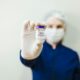 Na fotografiji se nalazi osoba u zaštitnoj medicinskoj odjeći koja nosi bijelu kiruršku masku i rukavice. Osoba drži u ruci malu bočicu botoxa s ljubičastim čepom, koju pokazuje prema kameri. Pozadina je jednostavna i svijetla, što dodatno naglašava bočicu u prvom planu. Fokus fotografije je na proizvodu, dok je lice osobe zamućeno, čime se stvara dojam profesionalnosti i čistoće medicinskog okruženja. Cjelokupni dojam slike sugerira kliničku preciznost i sigurnost u primjeni botoxa.