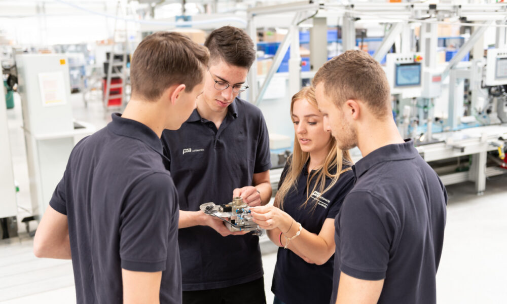 Četvero mladih ljudi u tamnoplavim majicama stoje u krugu u industrijskom pogonu i proučavaju tehnički uređaj koji drže u rukama.