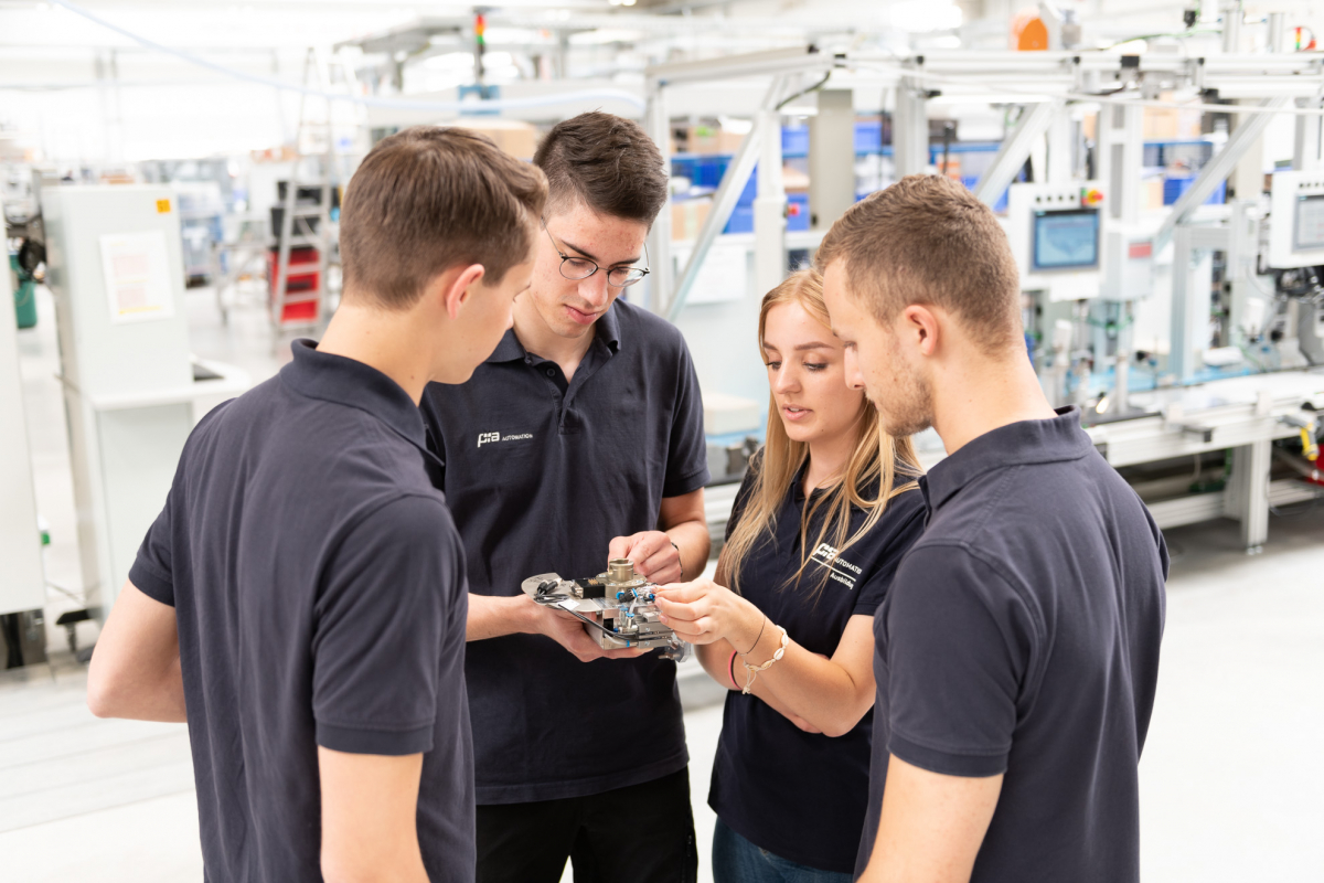 Četvero mladih ljudi u tamnoplavim majicama stoje u krugu u industrijskom pogonu i proučavaju tehnički uređaj koji drže u rukama.