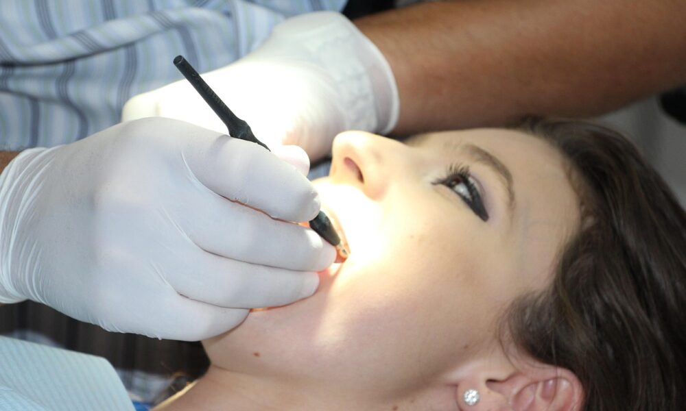 Na fotografiji je prikazan krupni kadar stomatološkog zahvata. Pacijentica leži zavaljena u stomatološkoj stolici, s otvorenim ustima, dok stomatolog ili dentalni higijeničar u bijelim rukavicama koristi dentalni instrument – vjerojatno zrcalo ili sondu – za pregled ili tretman zubi. Unutrašnjost usne šupljine osvijetljena je jakim svjetlom, što omogućuje precizan rad. Pacijentica nosi naušnice i ima šminku, što sugerira da se radi o rutinskom pregledu, a ne hitnoj intervenciji. Fotografija vizualno ističe važnost oralne higijene i profesionalne skrbi, uz dozu svakodnevne njege i povjerenja između pacijenta i stručnjaka.
