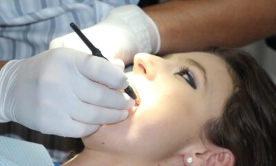 Na fotografiji je prikazan krupni kadar stomatološkog zahvata. Pacijentica leži zavaljena u stomatološkoj stolici, s otvorenim ustima, dok stomatolog ili dentalni higijeničar u bijelim rukavicama koristi dentalni instrument – vjerojatno zrcalo ili sondu – za pregled ili tretman zubi. Unutrašnjost usne šupljine osvijetljena je jakim svjetlom, što omogućuje precizan rad. Pacijentica nosi naušnice i ima šminku, što sugerira da se radi o rutinskom pregledu, a ne hitnoj intervenciji. Fotografija vizualno ističe važnost oralne higijene i profesionalne skrbi, uz dozu svakodnevne njege i povjerenja između pacijenta i stručnjaka.