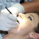 Na fotografiji je prikazan krupni kadar stomatološkog zahvata. Pacijentica leži zavaljena u stomatološkoj stolici, s otvorenim ustima, dok stomatolog ili dentalni higijeničar u bijelim rukavicama koristi dentalni instrument – vjerojatno zrcalo ili sondu – za pregled ili tretman zubi. Unutrašnjost usne šupljine osvijetljena je jakim svjetlom, što omogućuje precizan rad. Pacijentica nosi naušnice i ima šminku, što sugerira da se radi o rutinskom pregledu, a ne hitnoj intervenciji. Fotografija vizualno ističe važnost oralne higijene i profesionalne skrbi, uz dozu svakodnevne njege i povjerenja između pacijenta i stručnjaka.