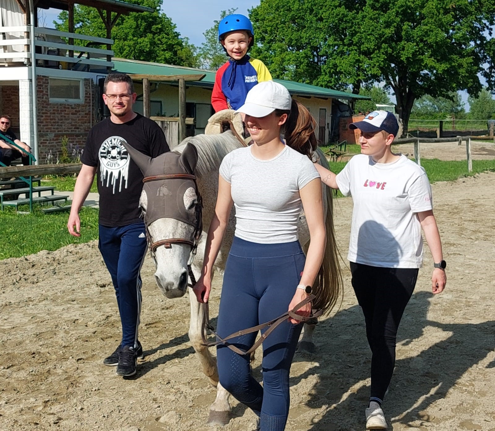 Na fotografiji se nalazi dijete koje jaše bijelog konja, okruženo s tri odrasle osobe koje hodaju uz njega. Dijete nosi plavi zaštitni kacigu i crveno-plavu odjeću te se smiješi dok sjedi na konju, što odaje dojam veselja i sigurnosti. Konj ima zaštitnu mrežicu preko glave (fly mask), a vodi ga žena u bijeloj šilterici i sivoj majici. Dvoje drugih odraslih – jedan u crnoj majici, drugi u bijeloj majici s natpisom "LOVE" – hodaju uz konja, vjerojatno pomažući i osiguravajući sigurnost djeteta.