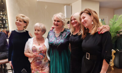 Pet nasmijanih žena pozira zagrljeno u elegantnom interijeru, odjevene u svečane haljine, s toplom i prijateljskom atmosferom.