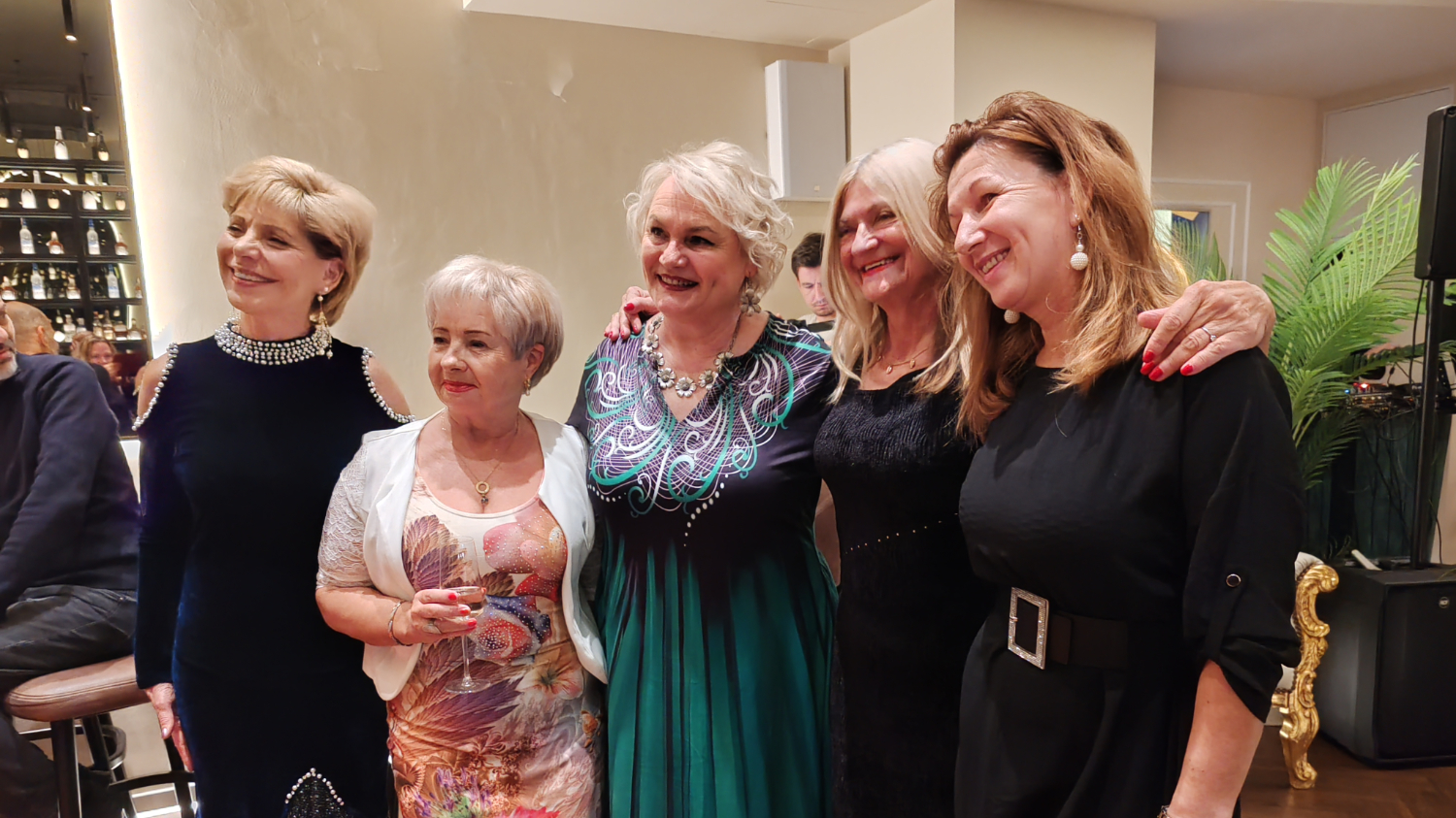 Pet nasmijanih žena pozira zagrljeno u elegantnom interijeru, odjevene u svečane haljine, s toplom i prijateljskom atmosferom.