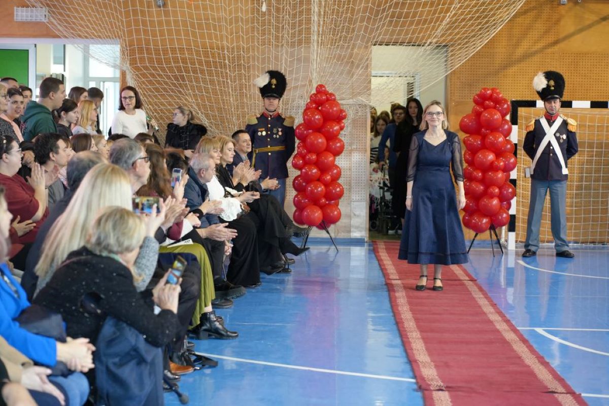 Fotografija prikazuje svečani trenutak u školskoj ili društvenoj sportskoj dvorani, pretvorenoj u ceremonijalni prostor. U središtu prizora, žena u elegantnoj tamnoplavoj haljini hoda niz crveni tepih, okružen s dvije strane stupovima od crvenih balona, što dodatno naglašava svečanost događaja. Na ulazu, s obje strane tepiha, stoje dvojica stražara u svečanim uniformama, mirno i dostojanstveno, dajući prizoru dojam formalnosti i poštovanja. S lijeve strane fotografije, publika sjedi na tribinama, a mnogi plješću, dok neki snimaju trenutak mobitelima, što sugerira da je dolazak žene važan dio programa. U pozadini, kroz ulaz, vidi se skupina ljudi, što može upućivati na to da je žena dio procesije, prezentacije ili dodjele priznanja. Pod dvorane je prepoznatljivo plave boje, a uočava se i sportska mreža, što potvrđuje da se radi o prenamijenjenom sportskom prostoru.