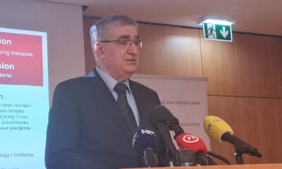 Muškarac u odijelu govori za govornicom ispred projekcijskog platna i mikrofona s oznakama televizijskih kuća tijekom stručnog skupa o genskim terapijama.