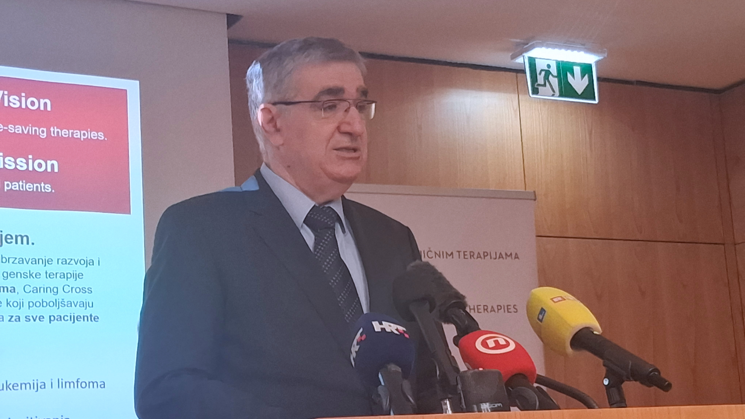 Muškarac u odijelu govori za govornicom ispred projekcijskog platna i mikrofona s oznakama televizijskih kuća tijekom stručnog skupa o genskim terapijama.