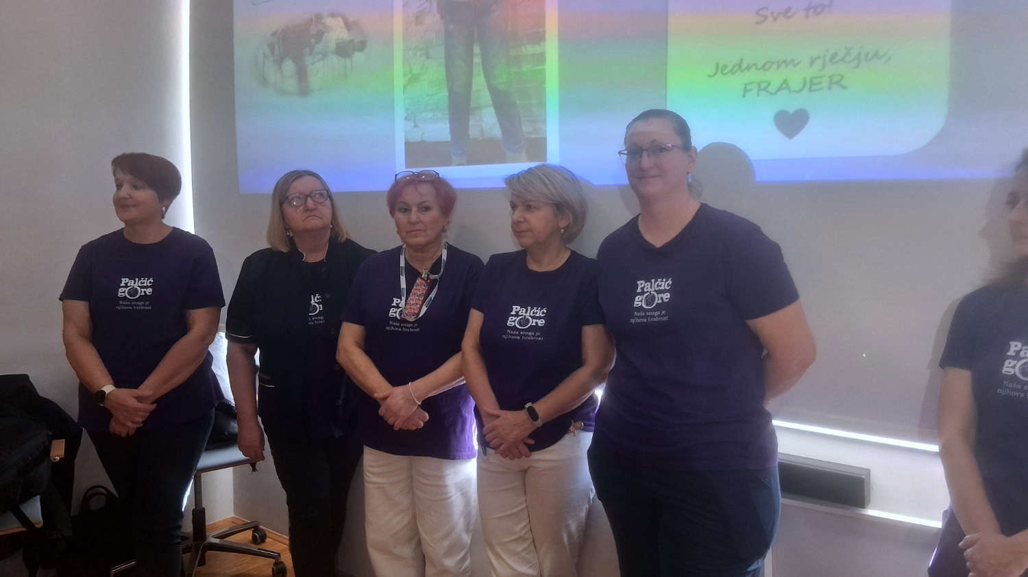 Skupina žena stoji u nizu ispred zida s projekcijom. Sve nose ljubičaste majice s natpisom ‘Palčić gore – Naša snaga je njihova hrabrost’. Gledaju prema publici, mirnog izraza lica.