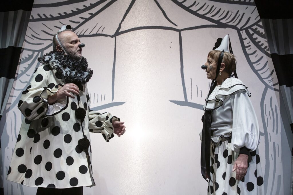 Dvoje glumaca u kazališnoj predstavi, odjeveni u crno-bijele kostime klaunova s velikim točkama, stoje jedno nasuprot drugome na pozornici. Oboje nose klaunske noseve i kapice, a iza njih je stilizirana crno-bijela scenografija.