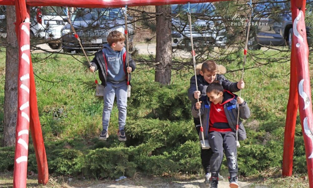 Na fotografiji su troje djece koja se igraju na ljuljačkama u vanjskom parku. Ljuljačke su crvene boje s bijelim ukrasnim uzorcima. Na jednoj ljuljački sjedi dijete u sivoj trenirci s natpisom "ENJOY P-LAY" na majici s kapuljačom. Na drugoj ljuljački zajedno se ljuljaju dvoje djece – jedno nosi crvenu Nike majicu s kapuljačom, a drugo tamnu jaknu s kapuljačom obrubljenom krznom. U pozadini se vidi zelena trava, grmlje, drveće i parkirani automobili, što stvara ugodnu atmosferu vanjskog prostora.