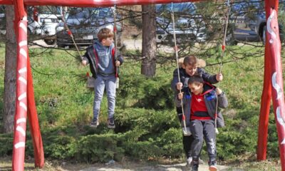 Na fotografiji su troje djece koja se igraju na ljuljačkama u vanjskom parku. Ljuljačke su crvene boje s bijelim ukrasnim uzorcima. Na jednoj ljuljački sjedi dijete u sivoj trenirci s natpisom "ENJOY P-LAY" na majici s kapuljačom. Na drugoj ljuljački zajedno se ljuljaju dvoje djece – jedno nosi crvenu Nike majicu s kapuljačom, a drugo tamnu jaknu s kapuljačom obrubljenom krznom. U pozadini se vidi zelena trava, grmlje, drveće i parkirani automobili, što stvara ugodnu atmosferu vanjskog prostora.
