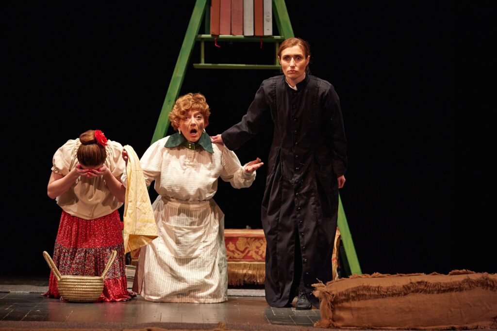 Tri glumca na kazališnoj pozornici u sceni iz predstave. S lijeva stoji glumica u narodnoj suknji i prekriva lice rukama, u sredini je glumica u svijetloj starinskoj haljini koja nešto objašnjava, a desno stoji glumac u tamnoj svećeničkoj odori. Iza njih je velika zelena drvena konstrukcija nalik ljestvama s knjigama.