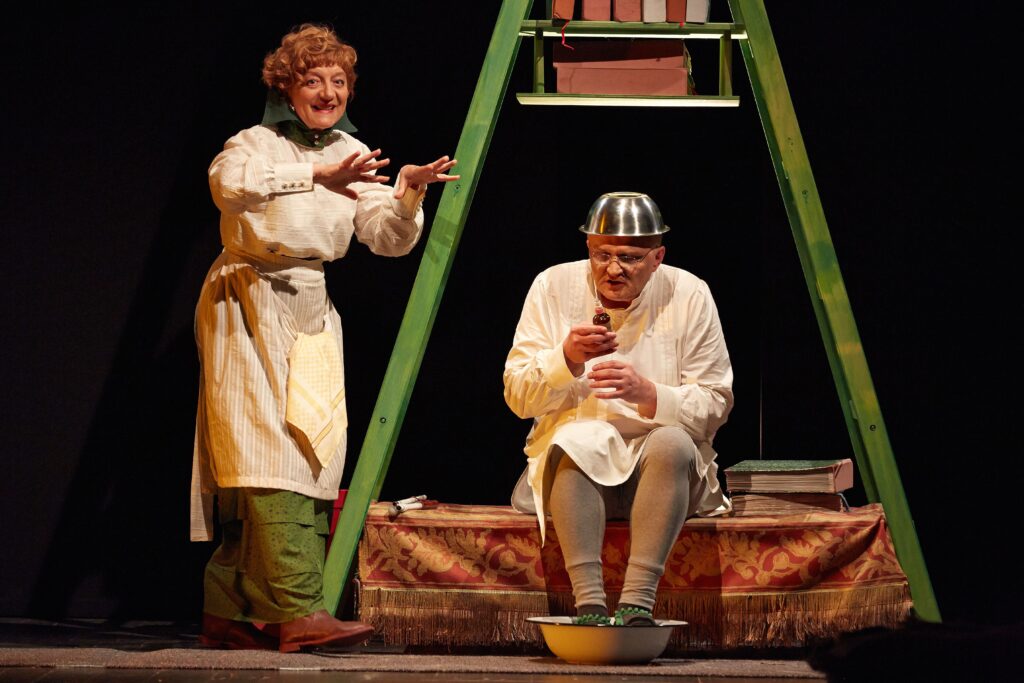 Glumica u svijetloj starinskoj haljini gestikulira prema glumcu koji sjedi i drži bočicu dok mu na glavi stoji metalna posuda. Ispred njega je lavor s vodom, a iza njih zelena konstrukcija s knjigama kao dio scenografije.