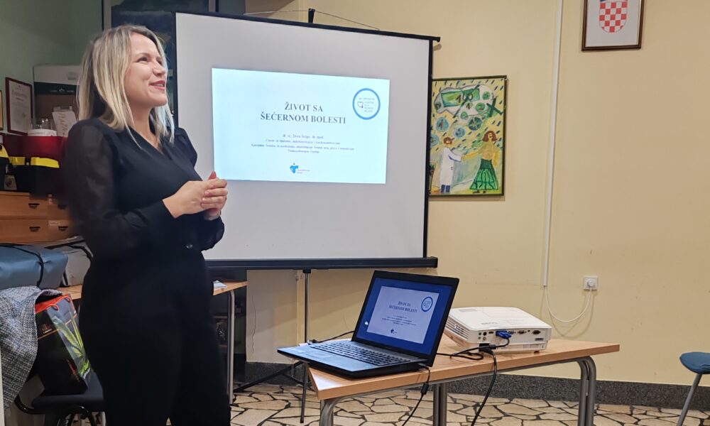Na slici se vidi žena koja stoji ispred projekcijskog platna i drži prezentaciju u prostoriji koja izgleda kao učionica ili dvorana za edukaciju. Na platnu i prijenosnom računalu prikazan je naslov prezentacije: "ŽIVOT SA ŠEĆERNOM BOLESTI" Prezentaciju drži dr. Daria Sorgo, dr. med