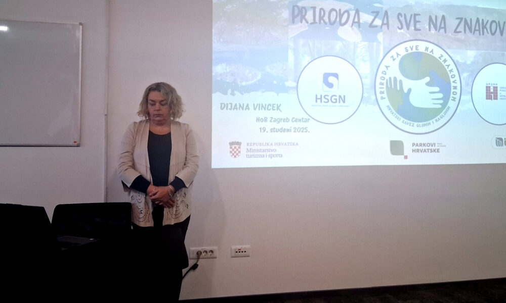 Žena stoji ispred projekcijskog platna tijekom prezentacije. Na platnu je prikazan naslov „Priroda za sve na znakovnom“, uz logotipe Hrvatskog saveza gluhih i nagluhih, Udruga osoba s invaliditetom, Parkovi Hrvatske i Ministarstva turizma i sporta. Žena ima svijetlu kosu, nosi bež jaknu preko tamne majice i stoji pokraj stola s laptopom.