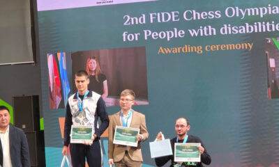 Fotografija prikazuje tri osobe na postolju s diplomama i medaljama. Iza njih na ekranu stoji natpis koji sugerira da se radi o drugoj olimpijadi u šahu za osobe s invaliditetom.