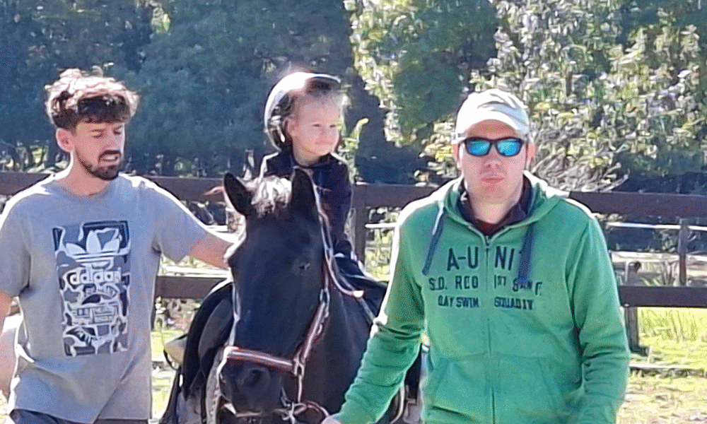 Dijete s kacigom sjedi na konju uz podršku dvojice odraslih muškaraca koji ga vode i pridržavaju tijekom jahanja u ograđenom prostoru, po sunčanom danu.