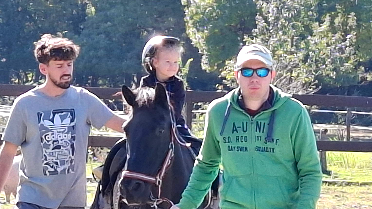 Dijete s kacigom sjedi na konju uz podršku dvojice odraslih muškaraca koji ga vode i pridržavaju tijekom jahanja u ograđenom prostoru, po sunčanom danu.