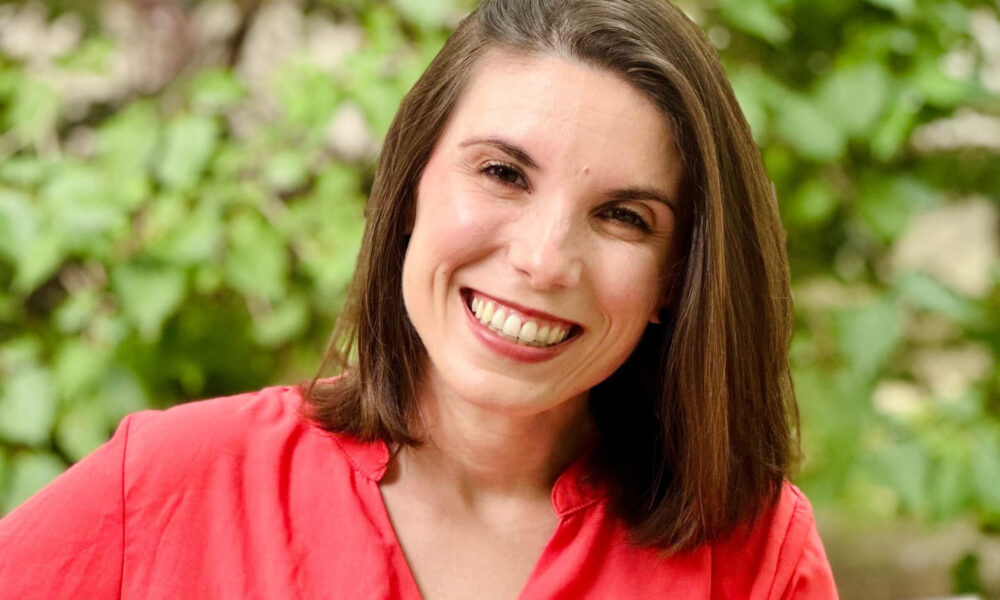 Fotografija prikazuje osobu koja sjedi na drvenoj klupi na otvorenom, okružena zelenim raslinjem u pozadini. Osoba nosi jarko crvenu bluzu i ima smeđu kosu do ramena. Smiješi se i gleda izravno u kameru, stvarajući dojam topline i pristupačnosti. Svjetlo je prirodno i ugodno, a ambijent djeluje mirno i prijateljski. Zbog skladne kompozicije i izražajnog izraza lica, fotografija je prikladna za osobnu ili profesionalnu upotrebu — primjerice kao portret, profilna slika ili promotivni vizual.