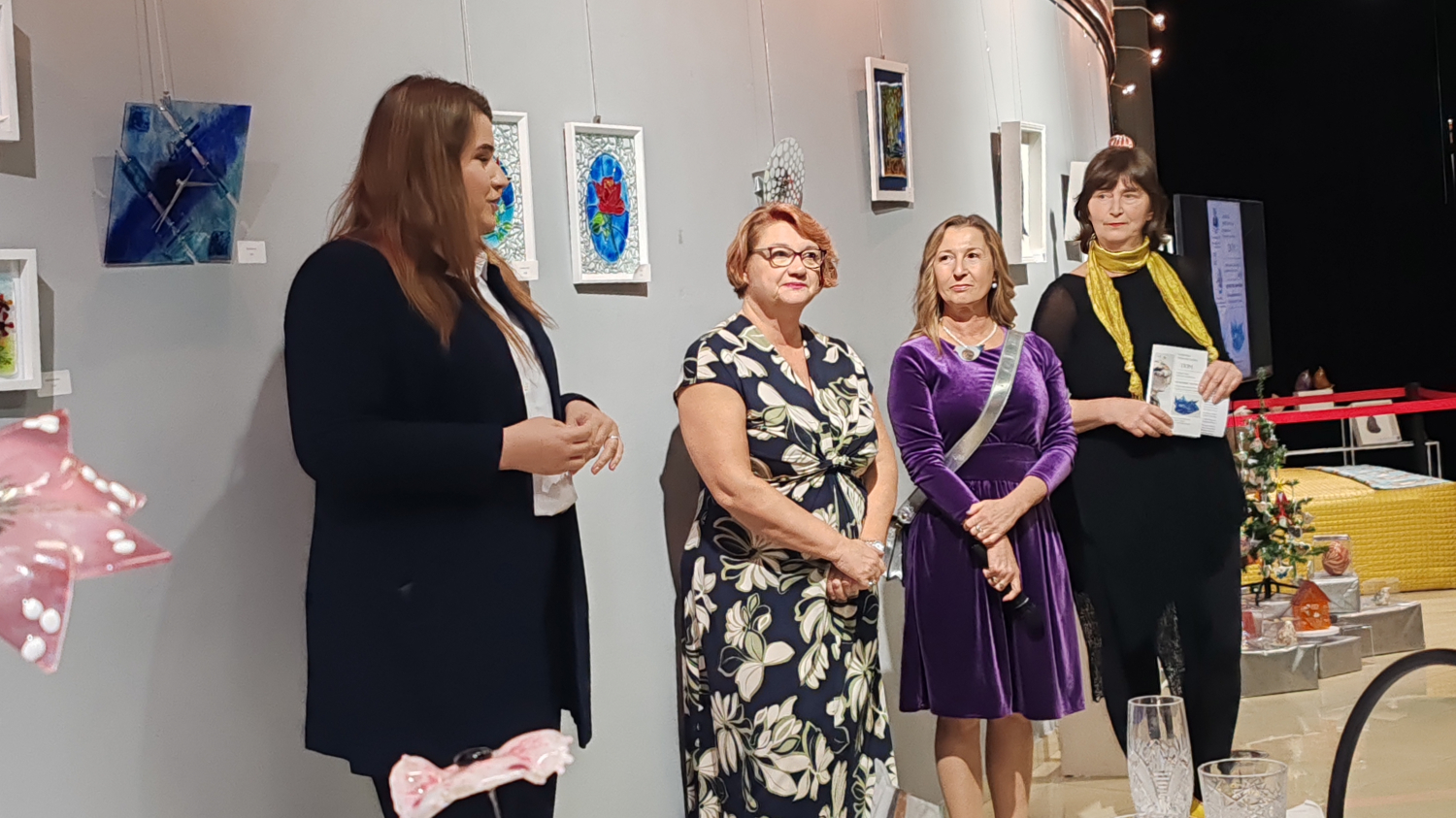 Četiri žene stoje uz izložene radove od stakla na zidu galerije, dok jedna od njih drži govor prilikom otvorenja izložbe.