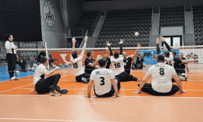 Fotografija prikazuje igrače koji igraju sjedeću odbojku u dvorani. Jedna je momčad u bijelim, a druga u tamnoplavim dresovima.