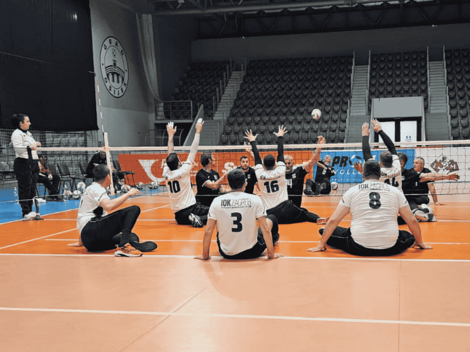 Fotografija prikazuje igrače koji igraju sjedeću odbojku u dvorani. Jedna je momčad u bijelim, a druga u tamnoplavim dresovima.