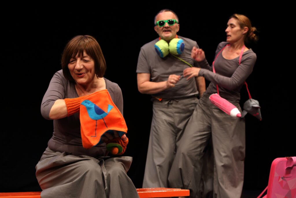 Tri glumca na pozornici nose siva kostimirana odijela u modernom, minimalističkom stilu. Glumica u prvom planu drži narančastu torbicu s motivom plave ptice, dok druga dva glumca iza nje u pokretu djeluju kao da sudjeluju u komičnoj sceni.