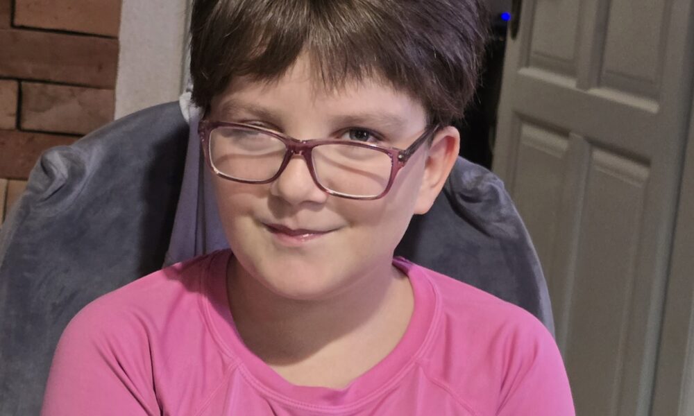 Na fotografiji je dijete kratke smeđe kose i naočala, koje sjedi u zatvorenom prostoru na sivoj stolici. Dijete nosi ružičastu majicu s natpisom „DANCE” ispisanim stiliziranim plavim i ružičastim slovima. U pozadini se vidi zid od cigle, bijela vrata i dio tamnog ormarića ili kućanskog aparata. Dijete ima izražaj na licu s jednim okom lagano zatvorenim i suptilnim osmijehom, što daje dojam spontanosti i osobnosti.