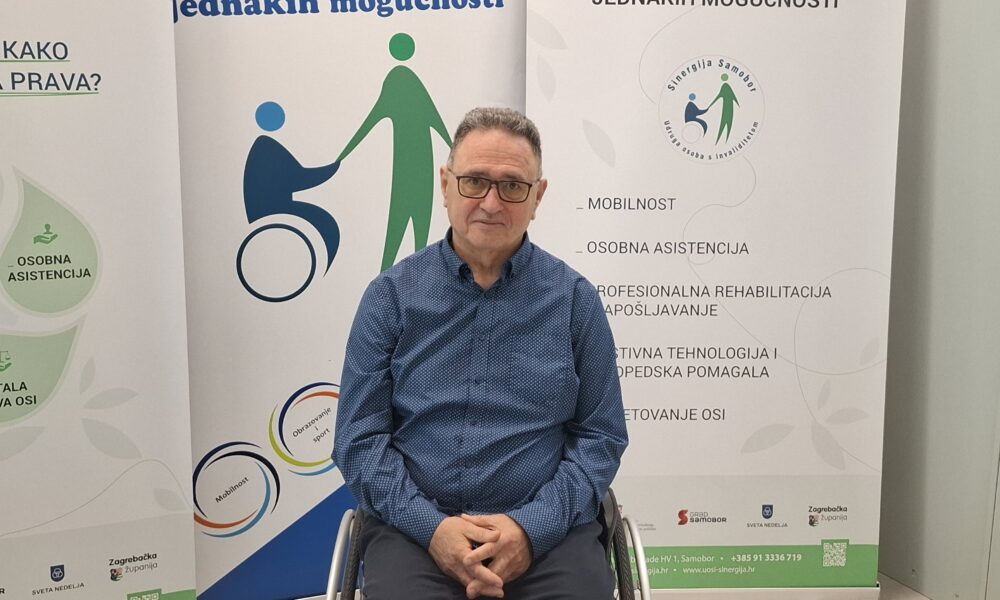 Fotografija prikazuje čovjeka u invalidskim kolicima, a iza njega se nalaze tri velika banera.