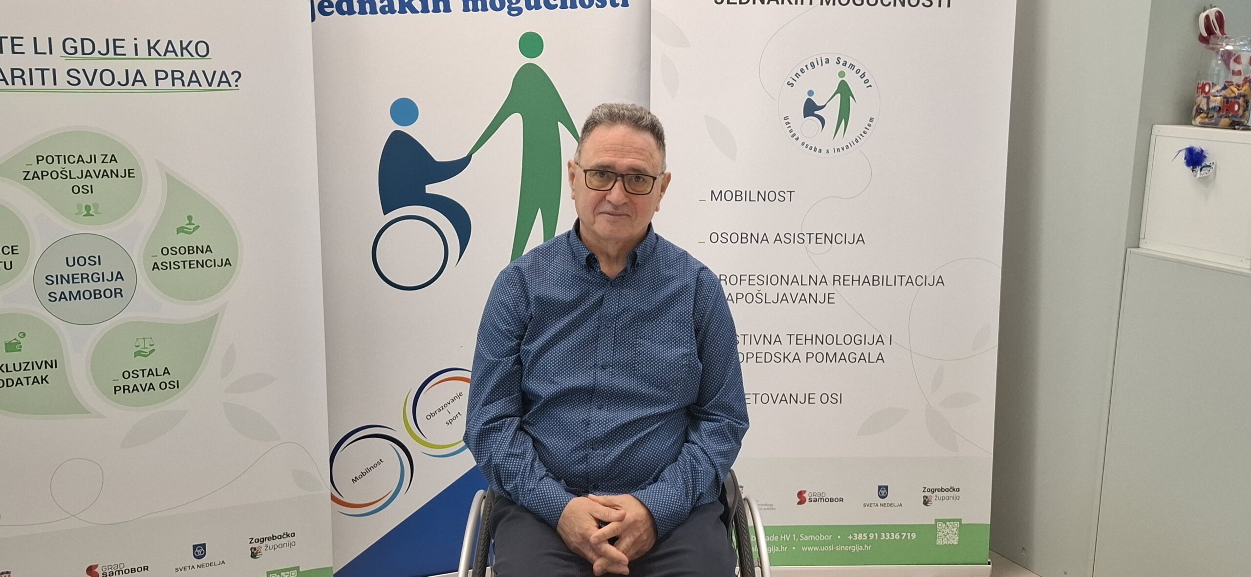 Fotografija prikazuje čovjeka u invalidskim kolicima, a iza njega se nalaze tri velika banera.
