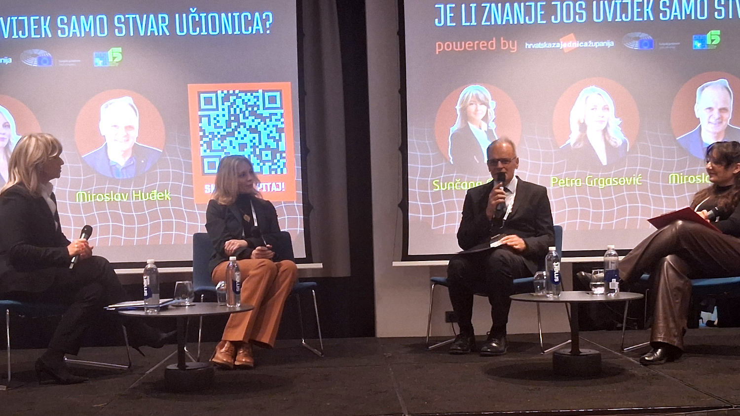 Četvero sudionika sjedi na pozornici tijekom panel-rasprave, ispred projekcije s naslovom „Je li znanje još uvijek samo stvar učionica?“ i prikazanim imenima govornika.
