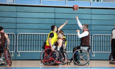 Fotografija prikazuje dvije ekipe koje igraju košarku u kolicima u dvorani. Jedna je ekipa u žutim dresovima, a druga u tamnocrvenim,