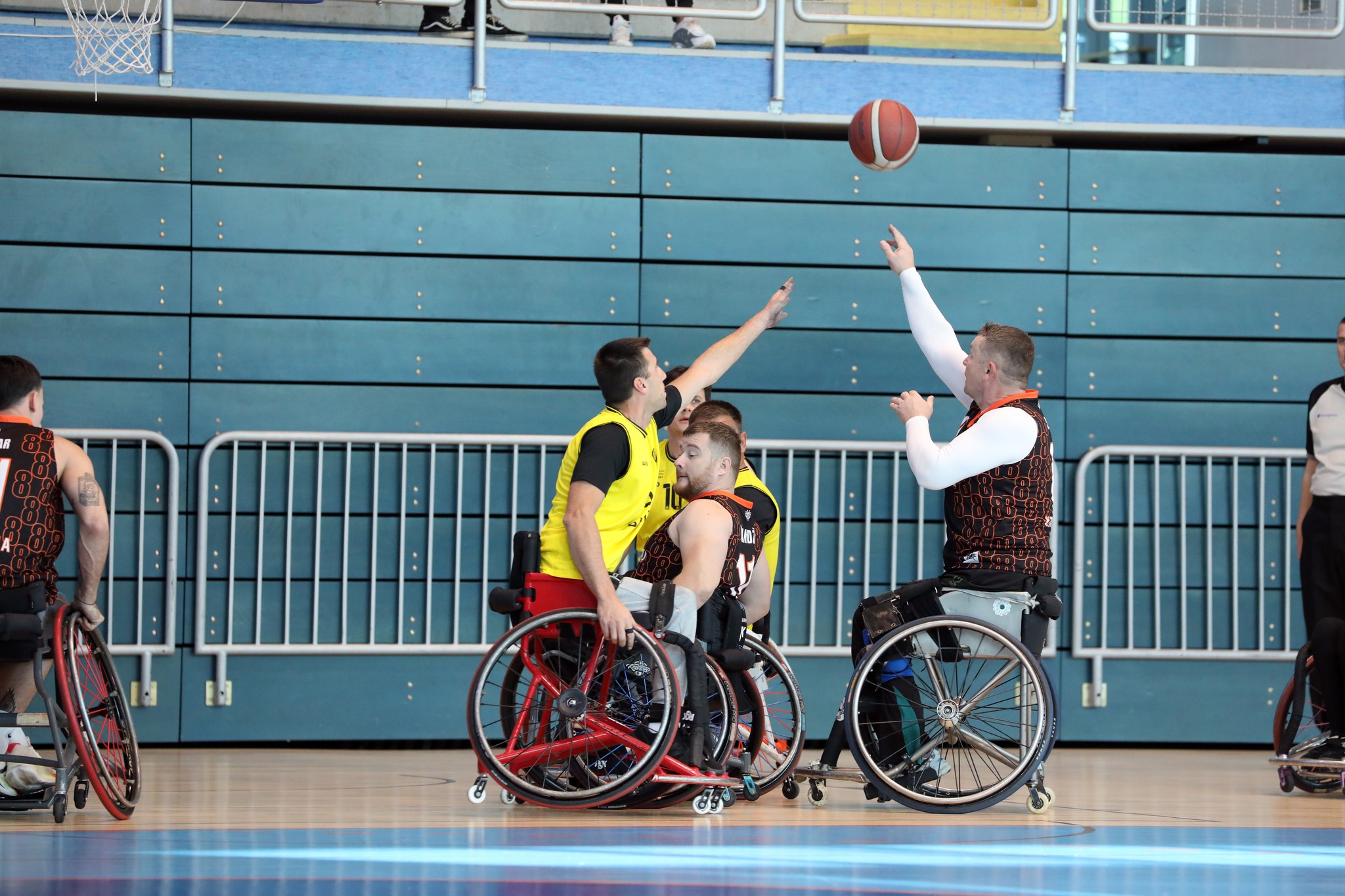 Fotografija prikazuje dvije ekipe koje igraju košarku u kolicima u dvorani. Jedna je ekipa u žutim dresovima, a druga u tamnocrvenim,