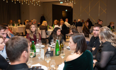 Fotografija prikazuje svečanu večeru ili banket u modernom, elegantno uređenom prostoru. U dvorani se nalaze brojni okrugli stolovi, svaki pažljivo postavljen s bijelim tanjurima, vinskim čašama i zelenim staklenim bocama vode. Gosti su odjeveni u poluformalnu do formalnu odjeću i međusobno razgovaraju, stvarajući ugodnu i živahnu atmosferu.