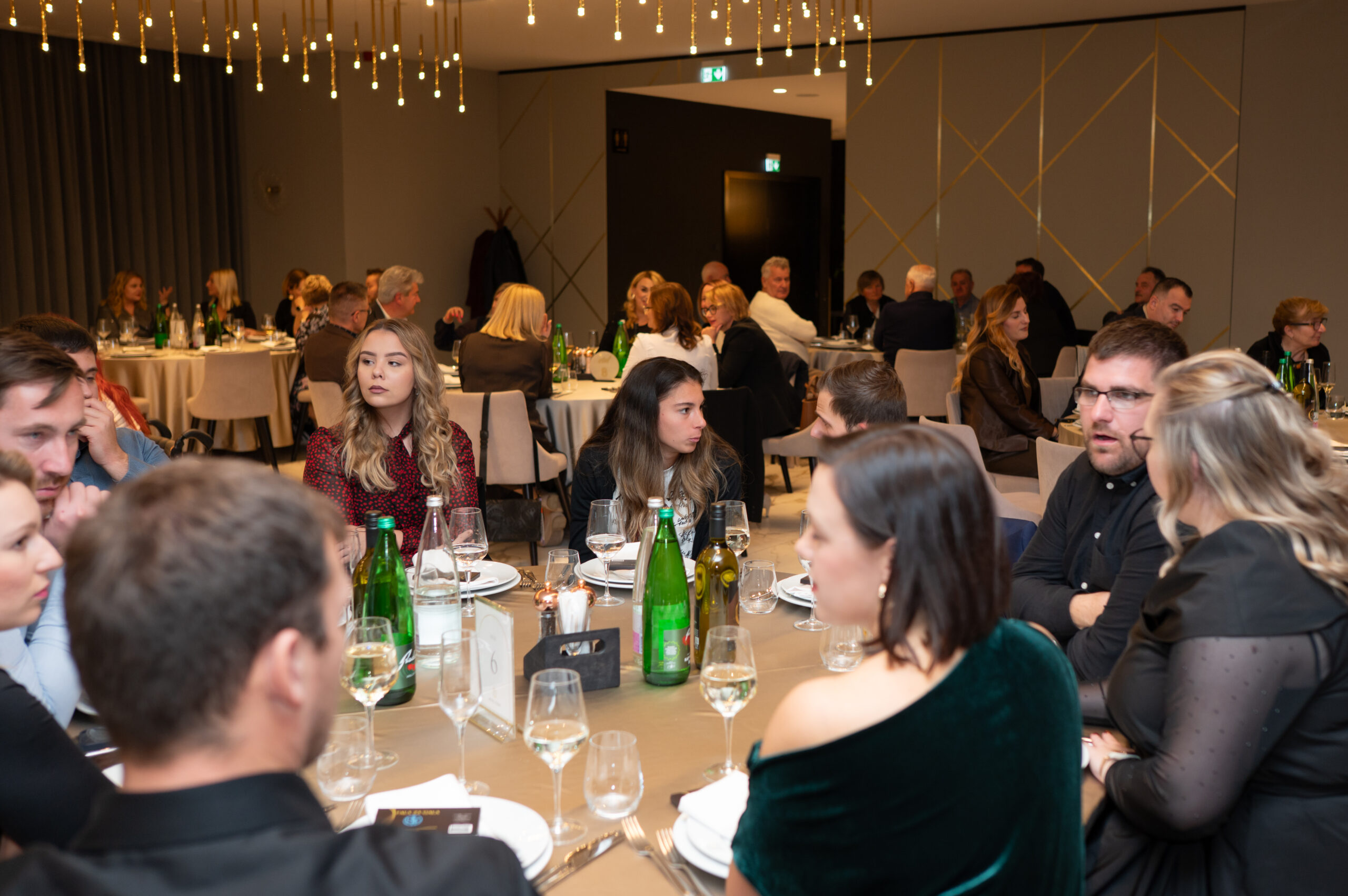 Fotografija prikazuje svečanu večeru ili banket u modernom, elegantno uređenom prostoru. U dvorani se nalaze brojni okrugli stolovi, svaki pažljivo postavljen s bijelim tanjurima, vinskim čašama i zelenim staklenim bocama vode. Gosti su odjeveni u poluformalnu do formalnu odjeću i međusobno razgovaraju, stvarajući ugodnu i živahnu atmosferu.