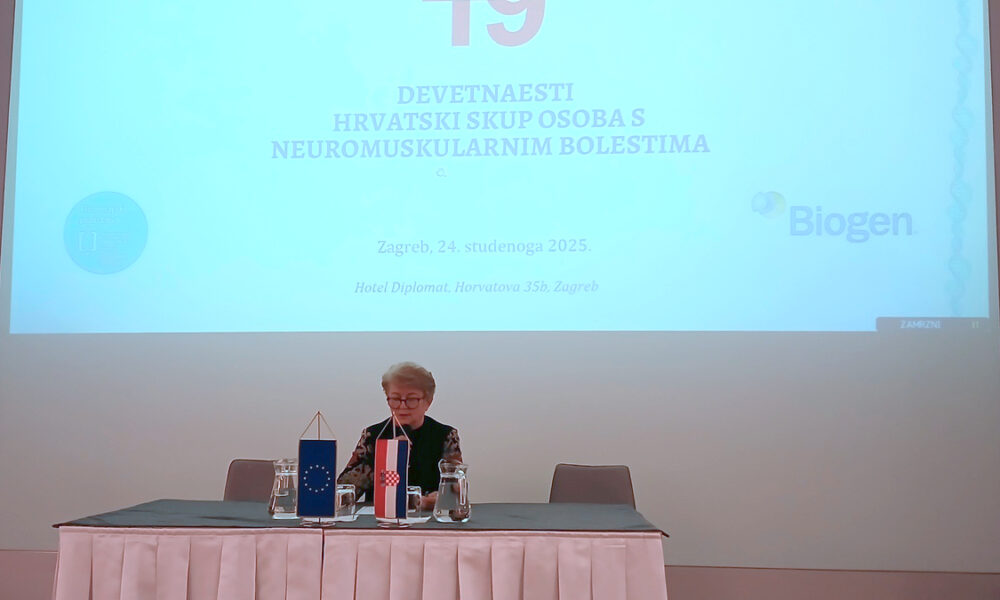 Na fotografiji se vidi konferencijska dvorana s velikim projekcijskim platnom na kojem piše “19. Devetnaesti hrvatski skup osoba s neuromuskularnim bolestima”, uz logotip Biogena. Ispred platna sjedi jedna osoba za stolom prekrivenim tamnom tkaninom, s dvije male stolne zastavice – europskom i hrvatskom. Na stolu su i čaše te staklene vrčevi s vodom. Prostorija je jednostavna i svijetla, a atmosfera je formalna i konferencijska.