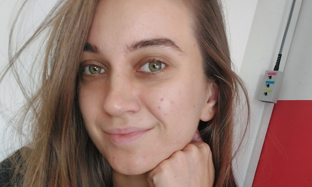 Portret mlade žene u krupnom planu. Ima dugu smeđukastu kosu, svijetlozelene oči i blagi osmijeh. Jednom rukom podupire bradu. Pozadina je jednostavna, u bijeloj i crvenoj boji, s malom električnom utičnicom i priključenim kabelom vidljivim u gornjem desnom kutu.