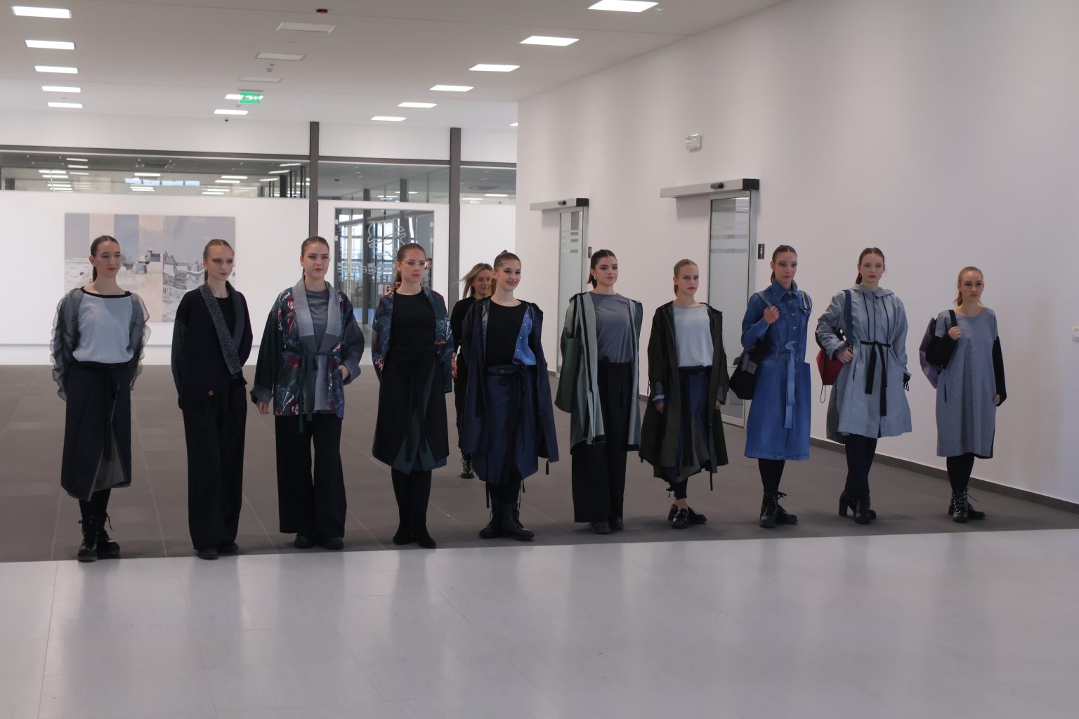 Na fotografiji je prikazana skupina od jedanaest osoba koje stoje u ravnoj liniji unutar modernog interijera. Prostor ima bijele zidove, jaku stropnu rasvjetu i minimalistički dizajn. Svi modeli nose usklađene, stilski dotjerane odjevne kombinacije koje djeluju kao dio modne kolekcije — moguće je da se radi o modnoj reviji ili prezentaciji. Odjeća je u tamnim i prigušenim tonovima, s naglaskom na slojevite komade poput dugih kaputa i čizama, stvarajući suvremen i skladan vizualni dojam. U pozadini se vide staklena vrata, hodnik i veliko umjetničko djelo na zidu, što dodatno doprinosi profesionalnoj i umjetničkoj atmosferi prostora. Cijela scena odiše elegancijom, kreativnošću i pažljivo osmišljenim stilom.