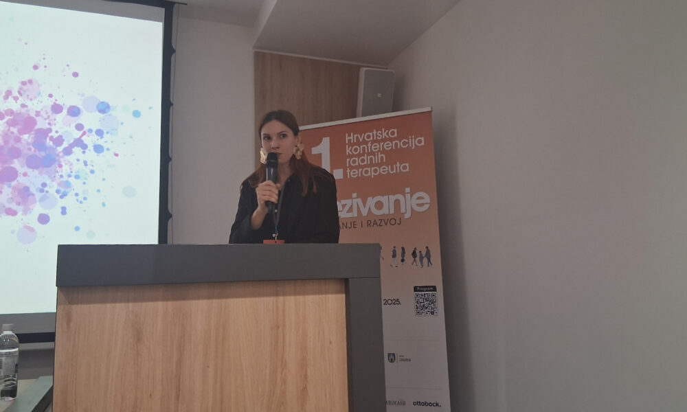 Žena stoji za govornicom dok drži mikrofon i govori na konferenciji. Obučena je u tamnu košulju i nosi velike zlatne naušnice. Iza nje je roll-up plakat narančaste boje s natpisom „1. Hrvatska konferencija radnih terapeuta“ i riječju „Izazivanje“. S lijeve strane vidi se dio projekcije s obojenim mrljama, a u prvom planu je drvena govornica. Na stolu pokraj projekcije stoji boca vode.