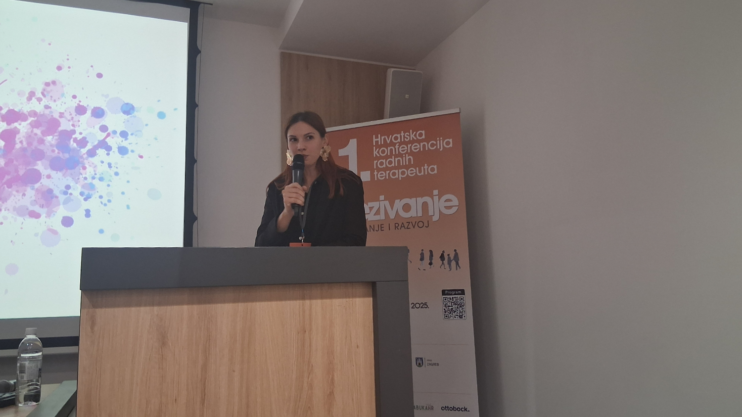 Žena stoji za govornicom dok drži mikrofon i govori na konferenciji. Obučena je u tamnu košulju i nosi velike zlatne naušnice. Iza nje je roll-up plakat narančaste boje s natpisom „1. Hrvatska konferencija radnih terapeuta“ i riječju „Izazivanje“. S lijeve strane vidi se dio projekcije s obojenim mrljama, a u prvom planu je drvena govornica. Na stolu pokraj projekcije stoji boca vode.
