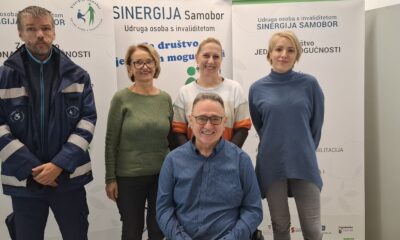 Fotografija prikazuje nekoiko ljudi od kojih je jedan čovjek u invalidskim kolicima. Iza njih su tri banera na kojima nešto piše.